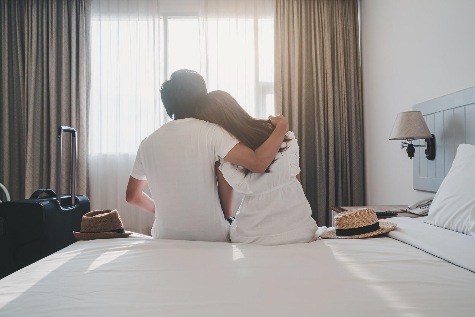Et par sidder med armen om hinanden på en opredt hotelseng med kufferten og to solhatte ved siden af sig