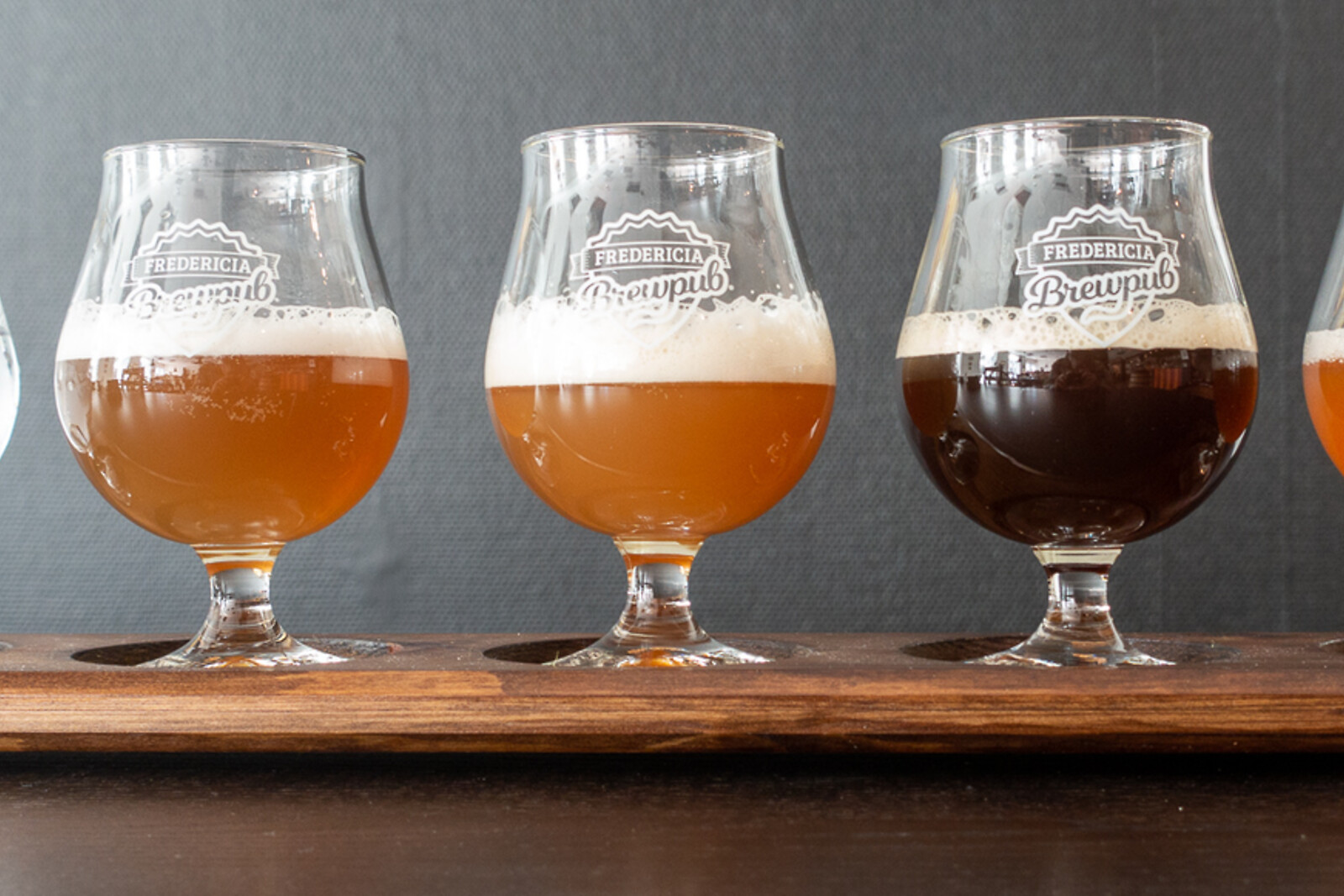 Tre ølglas med bryggeri-logo på et træbræt, fra lys gylden til mørk stout