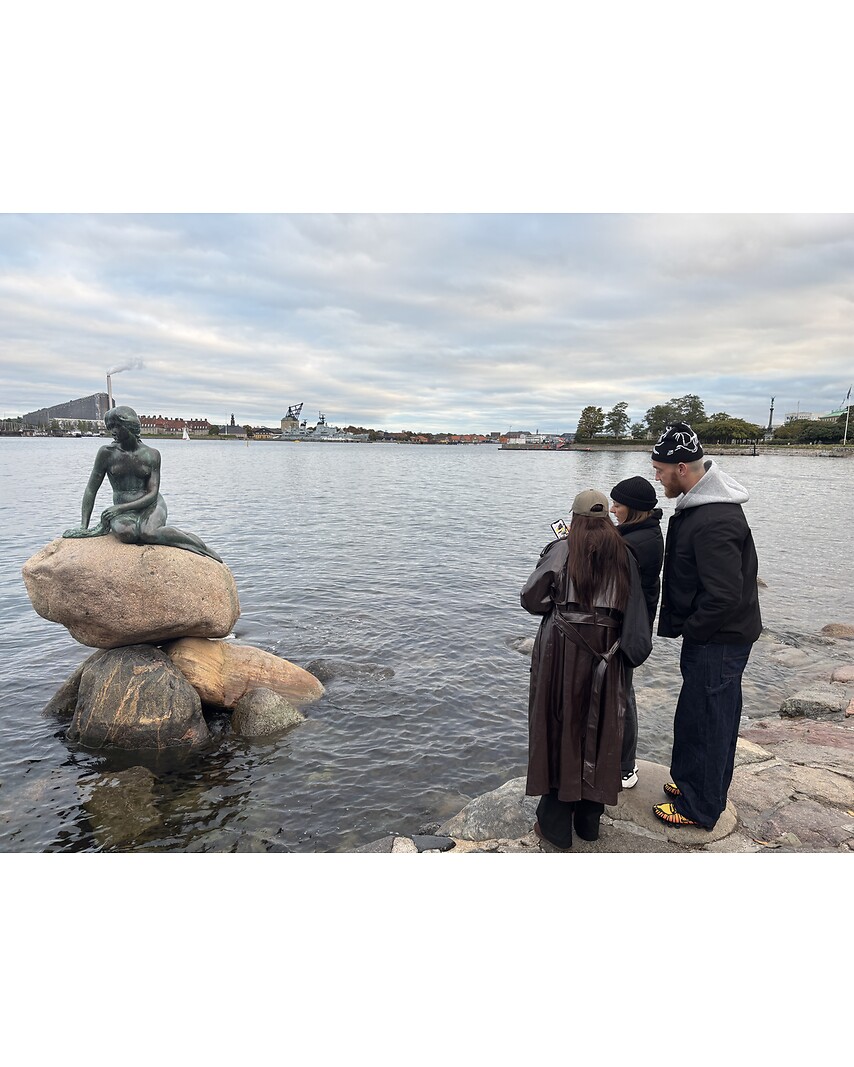 Tre personer i overtøj står ved Den Lille Havfrue og kigger på en seddel og en mobil